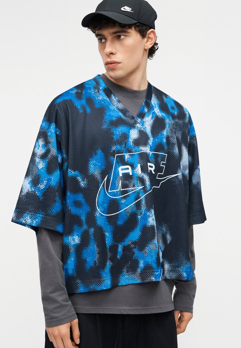 Junger Mann mit schwarzer Nike-Kappe und blau-schwarz gemustertem Nike Air Sporttrikot über grauem Langarmshirt, schaut nach links.