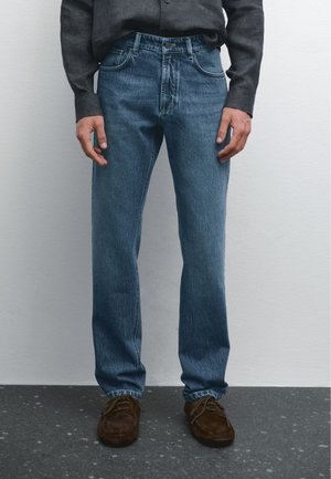 Blue denim jeans with a classic straight cut, five-pocket design, subtle whiskering, and a button fly, paired with brown suede shoes.