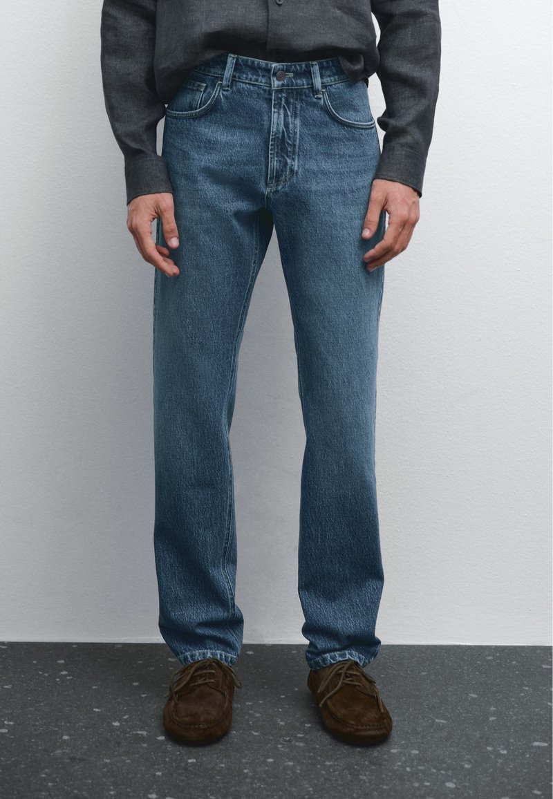 Blue denim jeans with a classic straight cut, five-pocket design, subtle whiskering, and a button fly, paired with brown suede shoes.