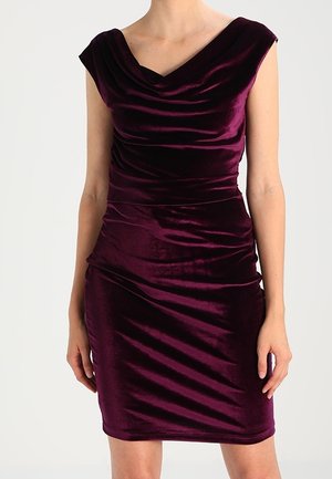 Kobieta w bezrękawną, głęboko purpurową welwetową sukienkę z drapowanym dekoltem i dopasowaną spódnicą za kolano na jednolitym tle.