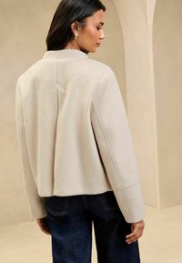 Lys beige, cropped jakke i uldblanding med høj hals, sømdetaljer og faldende skuldre, parret med mørke denimjeans.