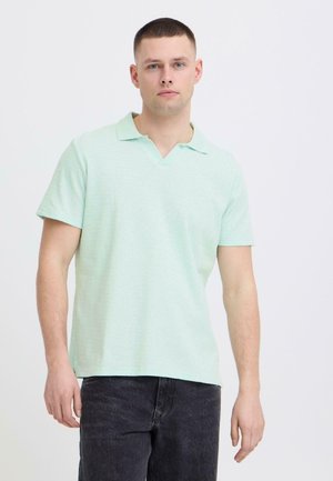 Jeune homme debout portant un polo à manches courtes vert menthe clair et un jean foncé, sur un fond blanc uni.