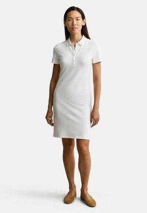Femme debout portant une robe polo blanche à manches courtes et des ballerines marron sur un fond gris clair uni.