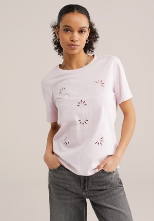 Kätega taskutes, esiküljele vaatav naine, kellel on lokkis juuksed, suurte rõngakujuliste kõrvarõngastega, heleroosa lillemustriga lõhikuga T-särk ja hallid teksad.