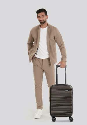 Schwarzer Hartschalenkoffer mit horizontalen Rillen, ausziehbarem Griff und drehbaren Rollen, positioniert neben einer Person in beiger Kleidung.