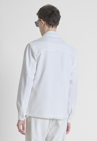 Camisa blanca de manga larga con puños abotonados. Presenta una textura sutil y un detalle de yugo en la parte posterior con un pequeño acento de hardware cerca del collar.