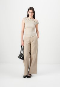 Beige, asimetrisks tops ar drapētiem elementiem, kombinēts ar gaiši beigiem plašiem biksēm; modele tur mazu melnu somiņu un valkā melnus zābakus.