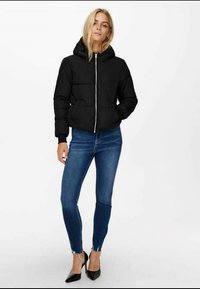 Black puffer jacket with a hood and front zipper, worn over fitted blue jeans with a frayed hem and black pointed-toe heels.