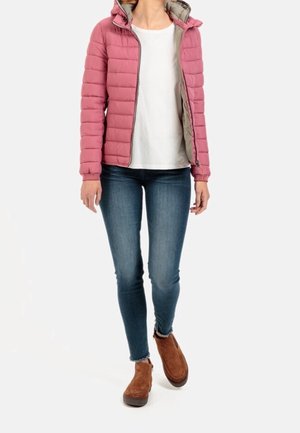 Kobieta w różowej pikowanej kurtce na białej koszuli, niebieskich obcisłych dżinsach i brązowych botkach, stojąca na białym tle.