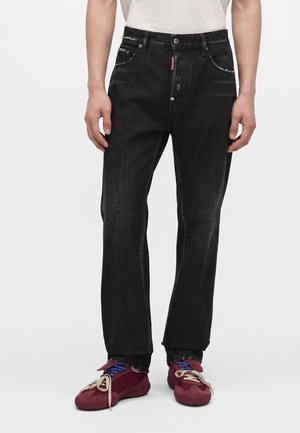 Personne portant un jean droit noir et des baskets bordeaux avec des lacets bleus et beiges, debout devant un fond uni.