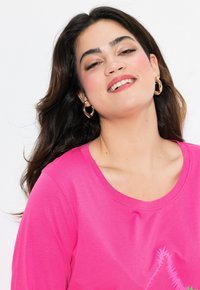 Camiseta de manga corta rosa con escote redondeado y un diseño texturizado en la parte frontal. El modelo tiene el cabello largo y oscuro, y lleva aros de oro.