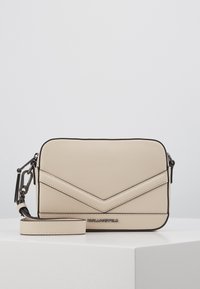 Beżowa torebka na ramię o fakturowanej powierzchni, z wzorem w jodełkę, czarną lamówką i odpinanym paskiem. Logo tłoczone z przodu.