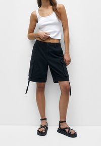 Pantalones cortos de carga negros con dos bolsillos laterales, una cremallera y cordones ajustables. Combinados con una camiseta corta blanca y sandalias negras.