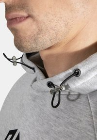 Lys grå hettegenser med metall-detaljer på snorene. Nærbilde som viser tekstur i stoffet, detalj på hetta og kontrasterende svart snor. Minimal merking synlig.
