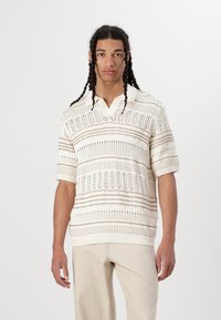 Polo tricot crème à manches courtes avec des motifs rayés texturés et un col en V, comportant des mailles ouvertes et des accents côtes.