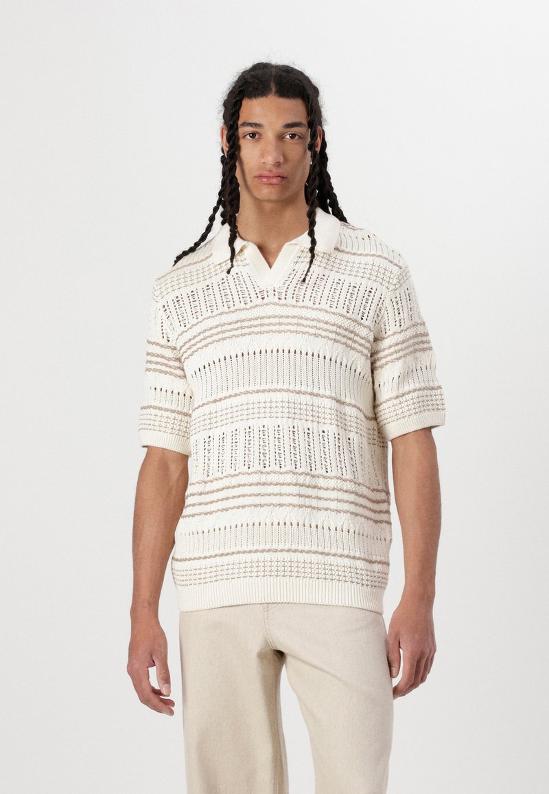 Polo tricot crème à manches courtes avec des motifs rayés texturés et un col en V, comportant des mailles ouvertes et des accents côtes.