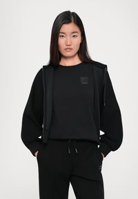 Zwarte sweatshirt met ronde hals, lange mouwen en een klein logo-patch, gecombineerd met bijpassende zwarte joggingbroek met trekkoord. Zachte stof en een relaxte pasvorm.