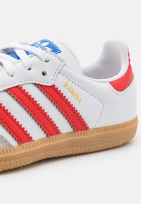Vita lädersneakers med röda ränder, gummisula, rund tå och ordet "SAMBA" i guld på sidan. Blå logotags på tungan.