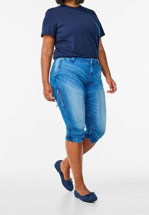 Kvinde iført blå denim capribukser med opkrogede manchetter, marineblå t-shirt og marineblå flade sko, stående med den ene fod let løftet.