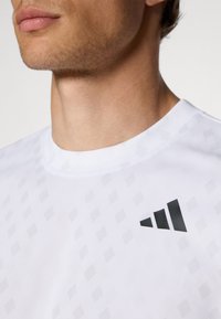 Weißes Sport-T-Shirt mit dezentem Diamantmuster, rundem Halsausschnitt und schwarzem Logo-Akzent auf der oberen linken Seite.