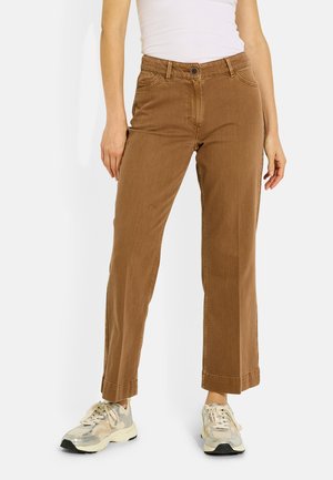 Mujer con pantalones marrones de pierna recta, camiseta sin mangas blanca y zapatillas beige, de pie frente a un fondo liso.