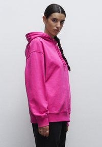 Světlá růžová mikina s kapucí, volného střihu, s přední klokaní kapsou a šňůrkou na kapuci. Hladká textilie.