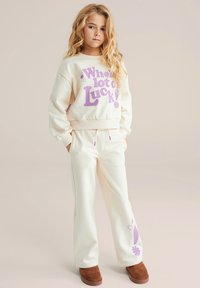 Kremna majica s kapuco s vijoličnim grafičnim tekstom; ujemajoče kremne hlače; rjavi semišni čevlji brez vezalk; mehka tkanina, sproščen kroj in pas z vrvico za zatezanje.