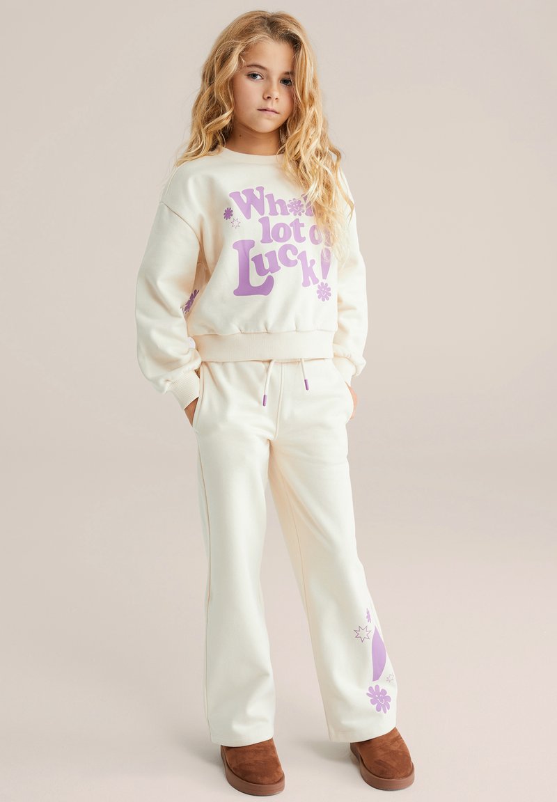 Kremna majica s kapuco s vijoličnim grafičnim tekstom; ujemajoče kremne hlače; rjavi semišni čevlji brez vezalk; mehka tkanina, sproščen kroj in pas z vrvico za zatezanje.