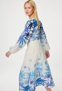 Langere mouw jurk met een lichtbeige stof en een blauwe tropische bloemenprint. Vloeiende rok, puffmouwen en elastische taille.