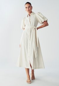 Mygtukų susegimas, dramblio kaulo spalvos lininė suknelė su pūstomis rankovėmis, juosmens raiščiu ir A formos sijonu; turi lygią tekstūrą ir kulkšnies ilgį.