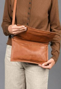 Bolso cruzado de cuero marrón con una textura suave, que cuenta con un cierre de solapa, herrajes plateados y un diseño delgado para un fácil transporte.