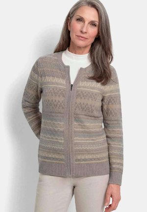 Frau mit langen grauen Haaren, die eine graue, gemusterte Reißverschluss-Strickjacke über einem weißen Top und hellen Hosen trägt, steht vor einem schlichten Hintergrund.