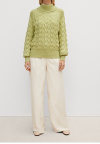 Hellgrüner Kabelstrickpullover mit hohem Kragen, gerippten Bündchen und Saum. Kombiniert mit cremfarbenen weitgeschnittenen Hosen und hellen Schuhen.