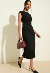 Schwarzes, figurbetontes Kleid mit Rundhalsausschnitt, seitlicher Raffung und goldenen Akzenten in der Taille. Model hält eine kleine burgunderfarbene Handtasche und trägt schwarze High Heels.