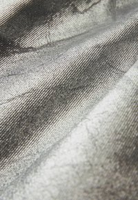 Metalická strieborná tkanina s textúrovaným povrchom. Tkanina je tesná, čo vytvára hladký lesk s jemnými vzormi viditeľnými v materiáli.