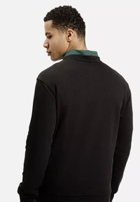 Czarny sweter z okrągłym kołnierzem, z teksturowanej tkaniny, z zieloną kratką widoczną przy szyi.