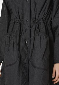 Sort quiltet yderjakke med snore i taljen, store frontlommer, trykknapper og struktureret stof. Har sorte pipings detaljer.