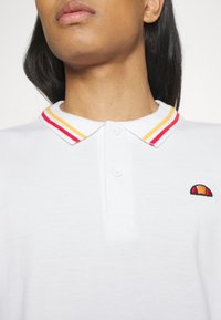 Biała koszulka polo z zapięciem na trzy guziki. Posiada kołnierzyk w czerwono-żółte paski oraz małe, wielokolorowe logo po lewej stronie.
