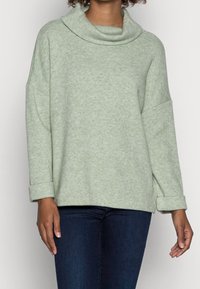 Pull-over à col bénitier en tissu doux vert clair, manches retroussées et coupe décontractée, assorti à un jean bleu foncé.