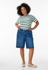 T-shirt rayé vert et blanc avec le texte "CASUAL", associé à un short en denim bleu moyen et des chaussures plates beige clair.