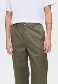 Olivegrüne Baumwollhose mit geradem Schnitt, ausgestattet mit einem vorderen Knopfverschluss und seitlichen Taschen. Glatte Textur, minimale Nähdetails.