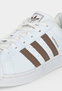 Άσπρο αθλητικό παπούτσι Adidas Superstar με καφέ ρίγες και κορδόνια, που δείχνει το λογότυπο της μάρκας στη γλώσσα και το κείμενο "SUPERSTAR" στο πλάι.