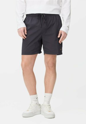 Mann trägt schwarze Shorts mit Kordelzug, weißes langärmeliges Oberteil, weiße Socken und weiße Turnschuhe, steht vor einem schlichten Hintergrund.