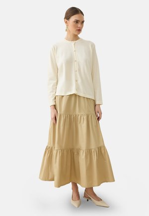 Femme portant un cardigan crème boutonné, une jupe longue beige à étages et des chaussures slingback beige à talon bas, debout devant un fond uni.