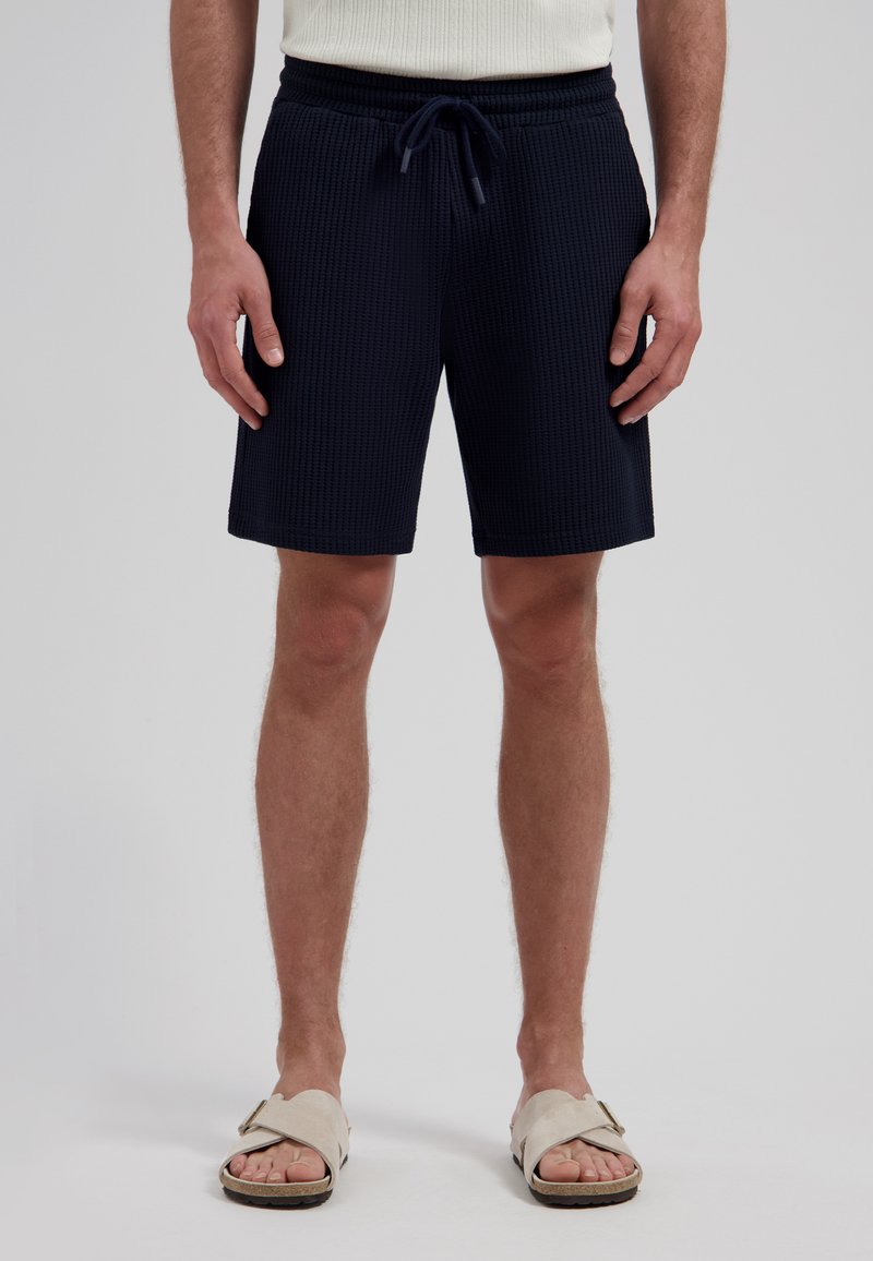Homme portant un short texturé noir avec cordon de serrage, un débardeur blanc et des sandales slides beiges, debout devant un fond uni.