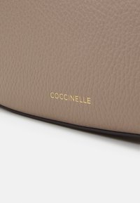 Texturiertes taupefarbenes Lederhandtasche mit einem goldenen "COCCINELLE"-Logo auf einer glatten Oberfläche, mit runder Form und kontrastierendem dunklen Rand.