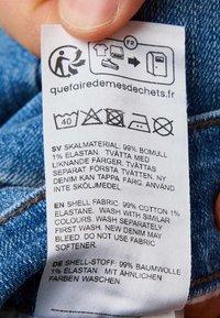 Etykieta ubrania przedstawiająca symbole prania, skład tkaniny: 99% bawełny, 1% elastanu oraz instrukcje pielęgnacji w języku szwedzkim, angielskim i niemieckim.