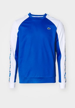 Zils džemperis ar baltām raglanu piedurknēm. Uz krūtīm ir zaļš Lacoste logo un uz piedurknes ir teksts. Izgatavots no mīksta materiāla.