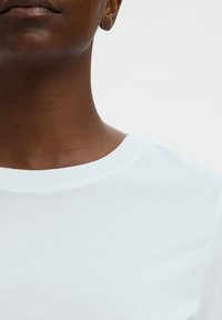Primo piano del collo e della parte inferiore del viso di una persona che indossa una maglietta bianca semplice con scollo tondo su sfondo bianco.