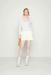 Top checkered lilás claro com mangas volumosas, calções brancos e sapatos de plataforma beiges. Segurando uma bolsa de madeira trançada. Fundo simples.